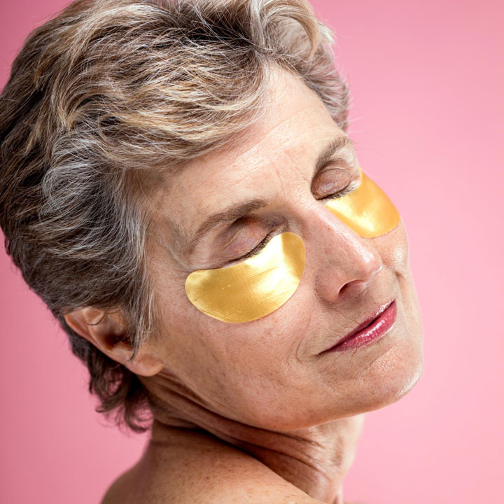 Older woman with under-eye patches smiling during a skincare routine focused on hydration and rejuvenation.